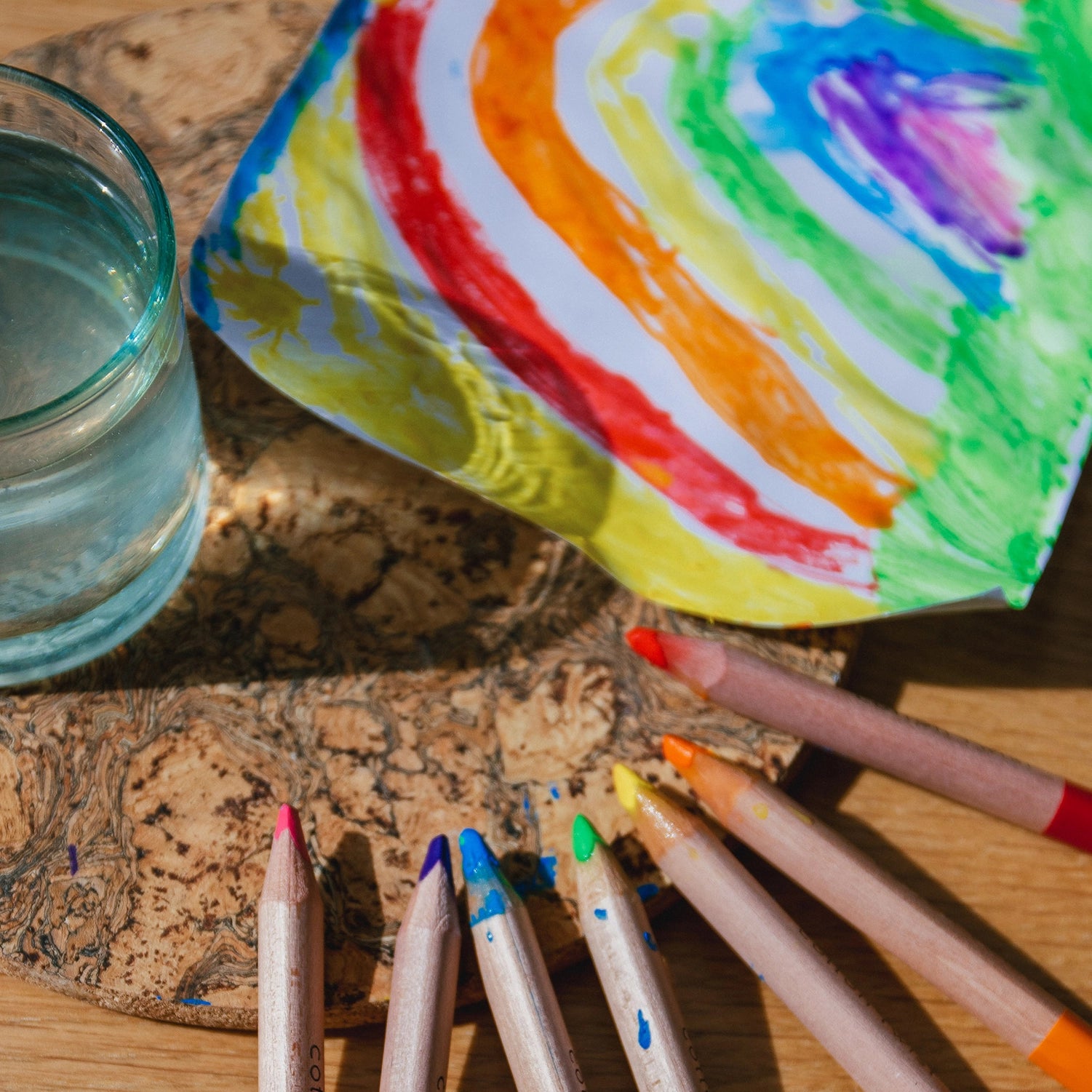 Colorful crayons and a glass of water on a wooden surface with a rainbow-colored paper in the background.