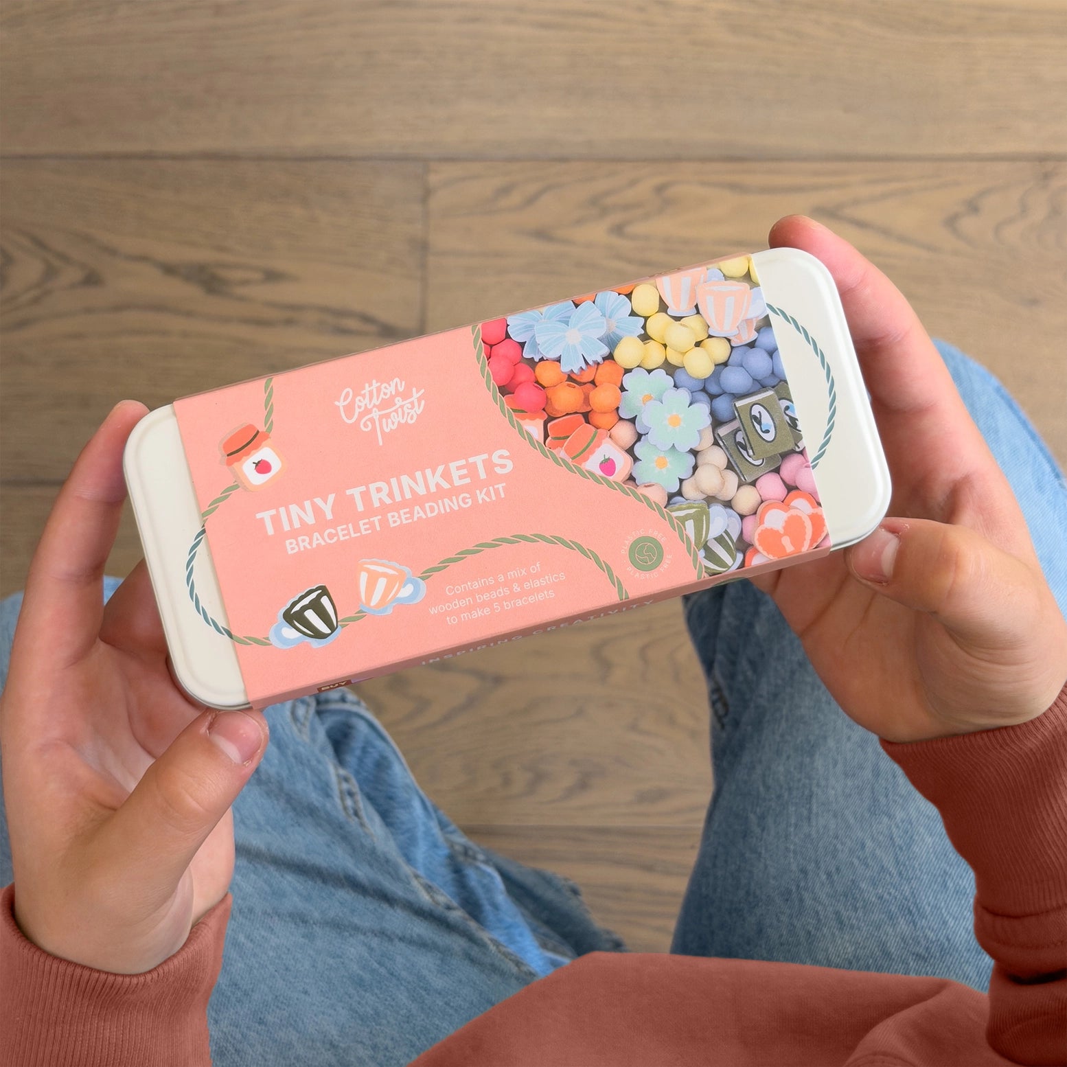 Person holding a 'Tiny Trinkets' bracelet beading kit packaging on a wooden floor.