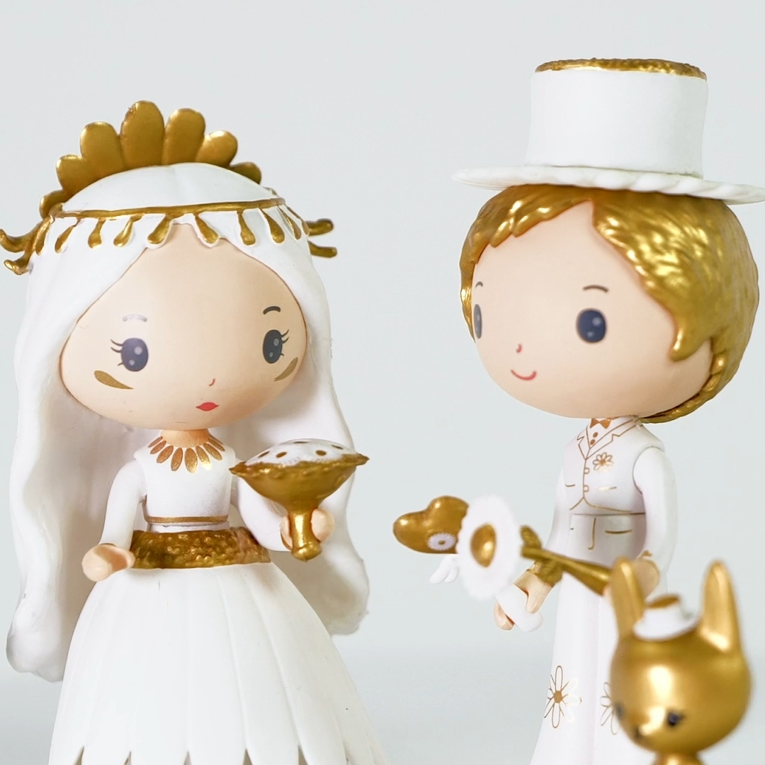 Two figurines, one in a white and gold dress with a crown, the other in a white outfit with a hat, on a light gray background.