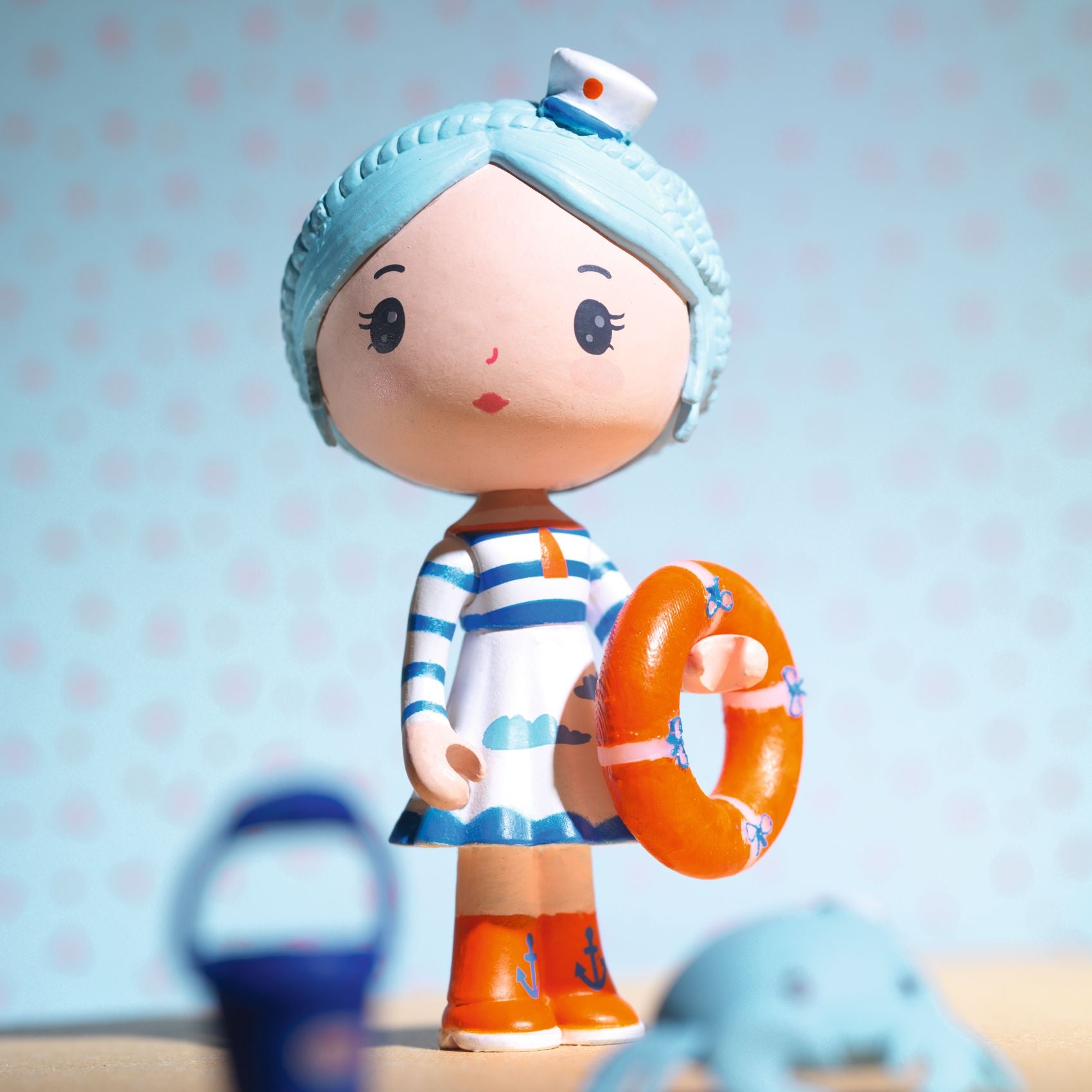 Toy figure with a blue hat and striped shirt holding an orange life buoy against a light blue background.