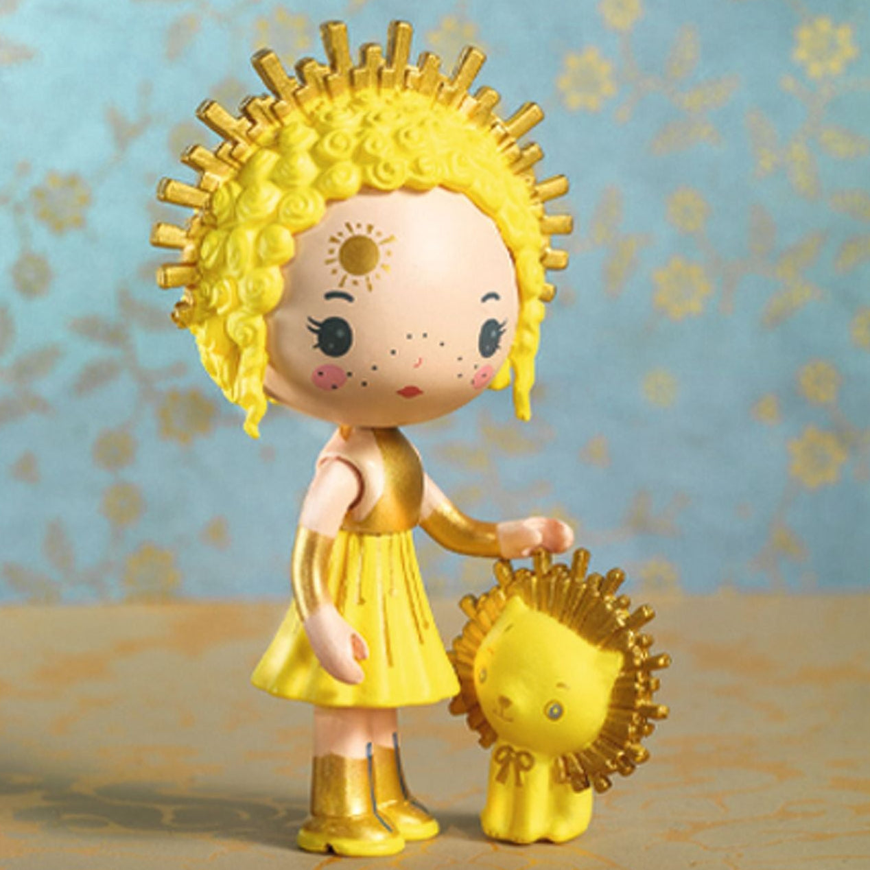 Toy figure of a child with yellow hair and dress standing with a small yellow lion against a floral-patterned background