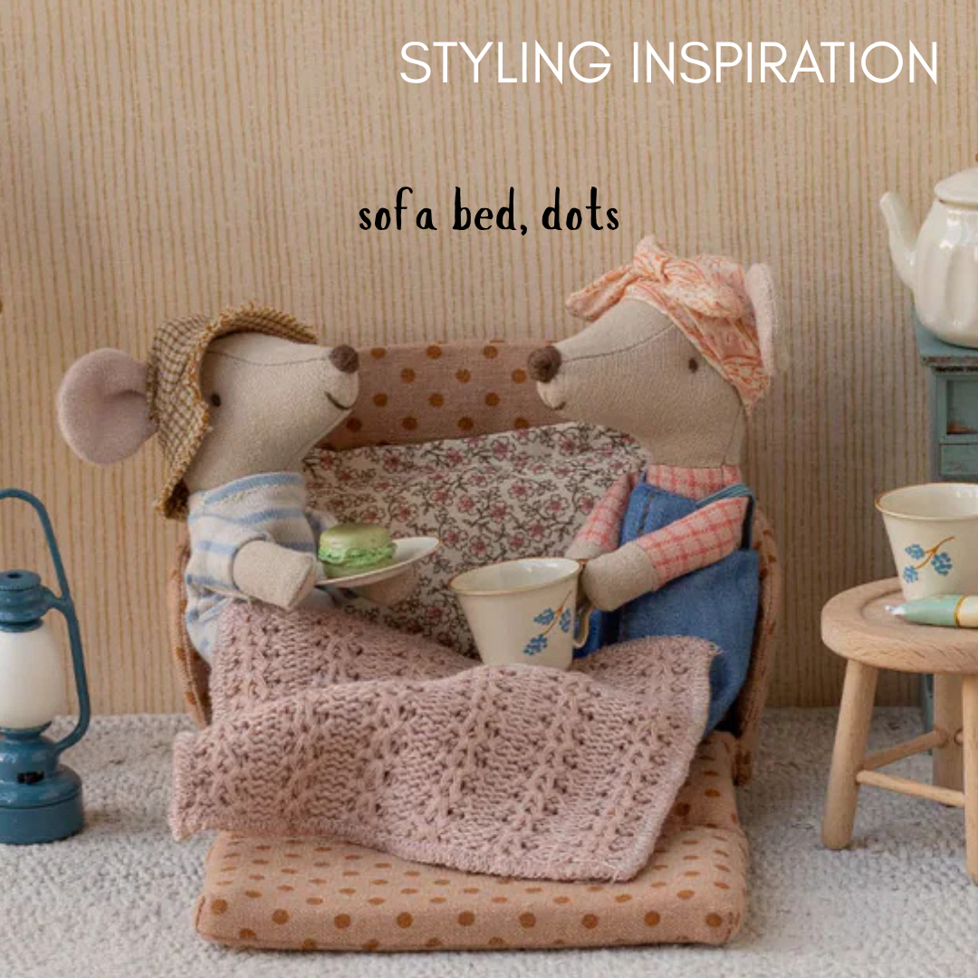 Two toy mice sitting on a miniature sofa with a polka dot blanket, surrounded by small furniture and items.