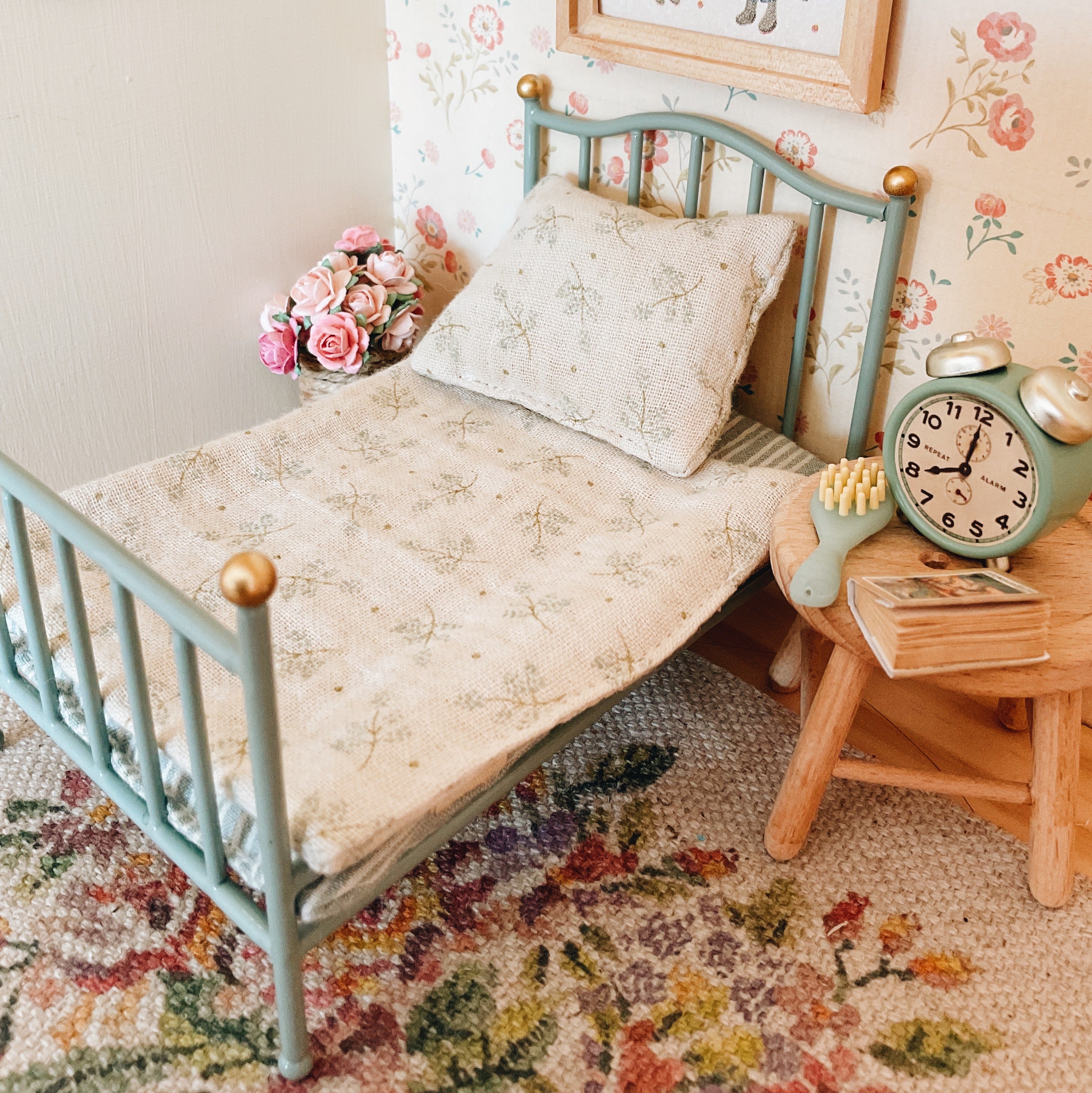 A Maileg miniature bed with a light blue metal frame, dressed in a floral-patterned quilt and matching pillow, styled in a vintage room scene with a wooden side table, alarm clock, and wallpaper.