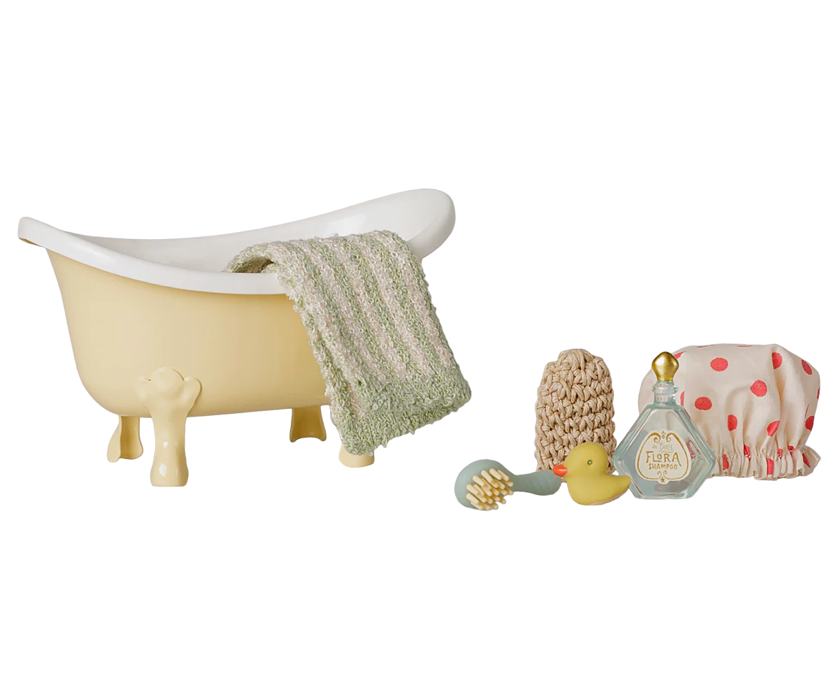 A miniature dollhouse-style bathroom set featuring a small cream bathtub with a green-and-white striped towel draped over the side, plus matching accessories including a yellow rubber duck, beige scrubber, blue brush, and a pink polka-dot bath mat.