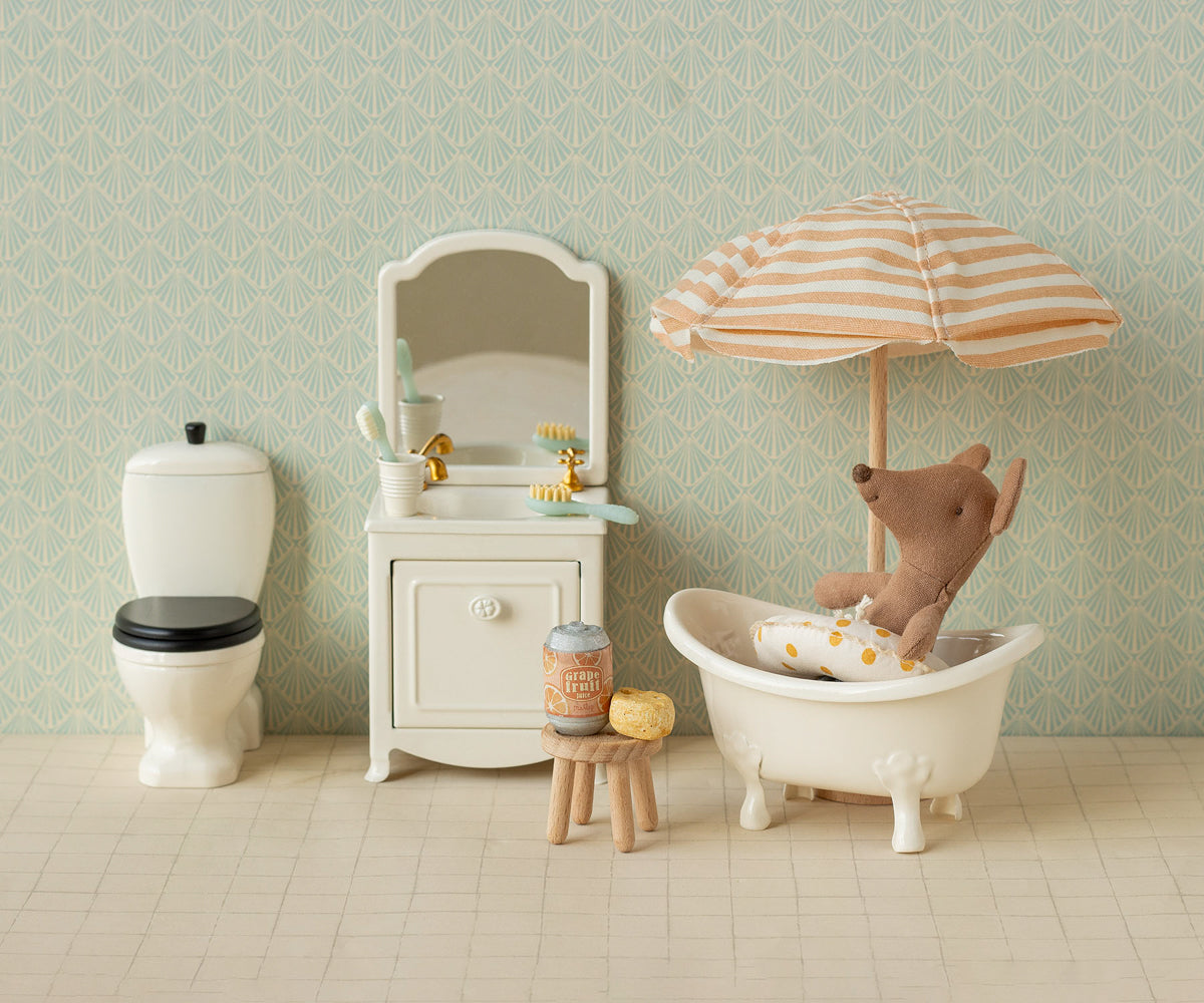 Miniature bathroom set with bathtub, toilet, and teddy bear under an umbrella against a patterned wall.