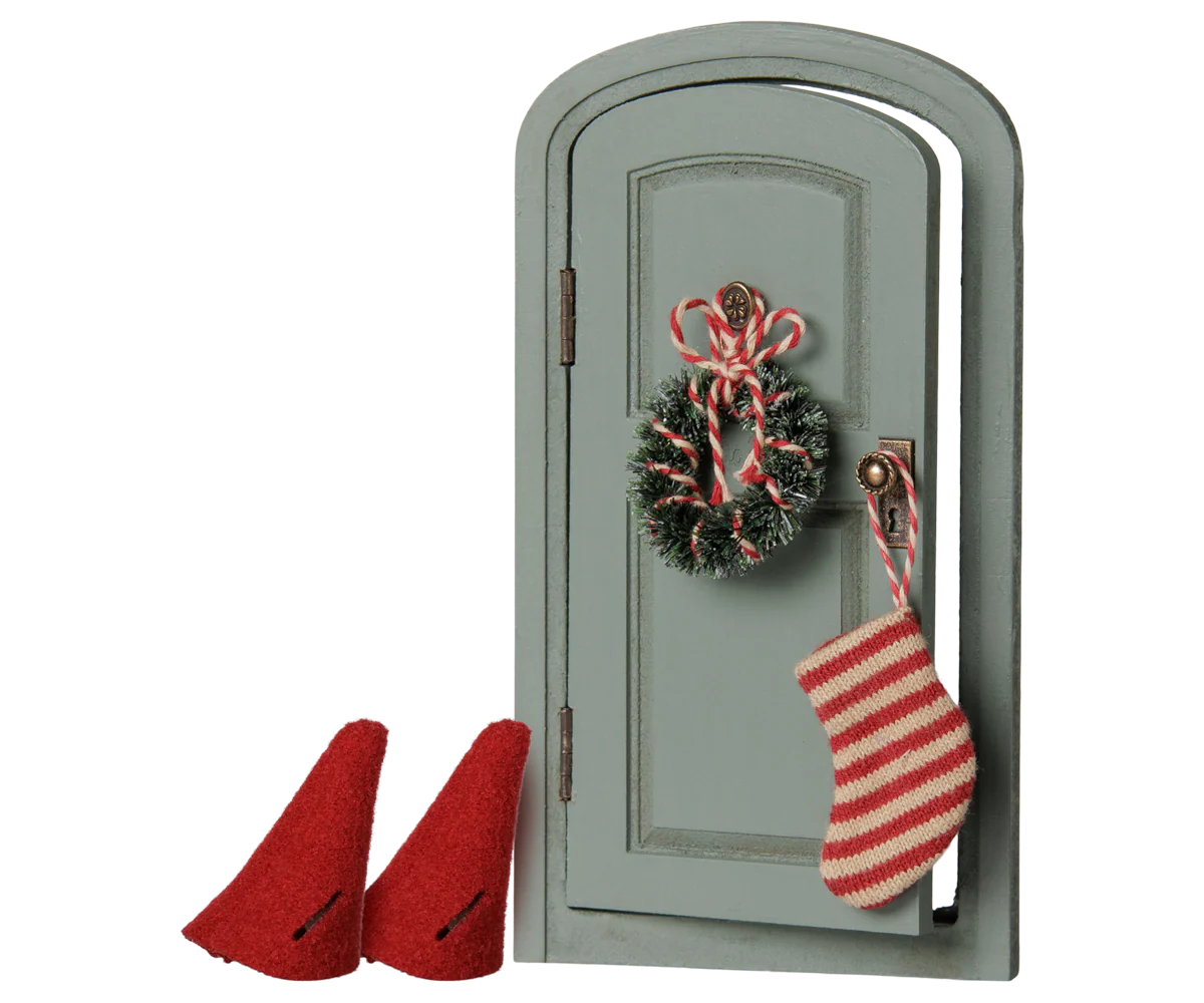 A miniature-style gray arched door decoration with a red-and-white striped stocking hanging from the handle and a matching wreath ornament, shown on a plain white background with two small red boot shapes at the lower left.