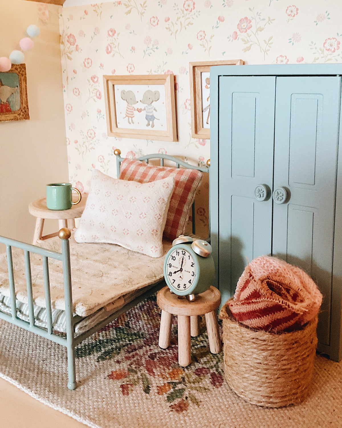 A styled miniature Maileg dollhouse bedroom scene featuring a light blue vintage bed with a patterned quilt and pillow, a matching blue closet, a small wooden side table with a mint green mug, a vintage-style green alarm clock on a stool, and a woven basket holding a pink knit blanket.