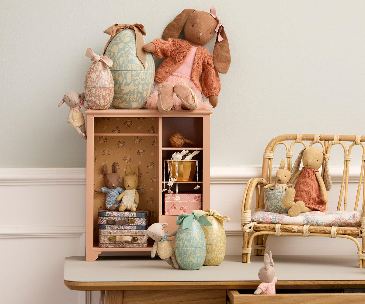 Collection of plush toys and maileg metal easter egg tins on a wooden shelf and chair against a light-colored wall.