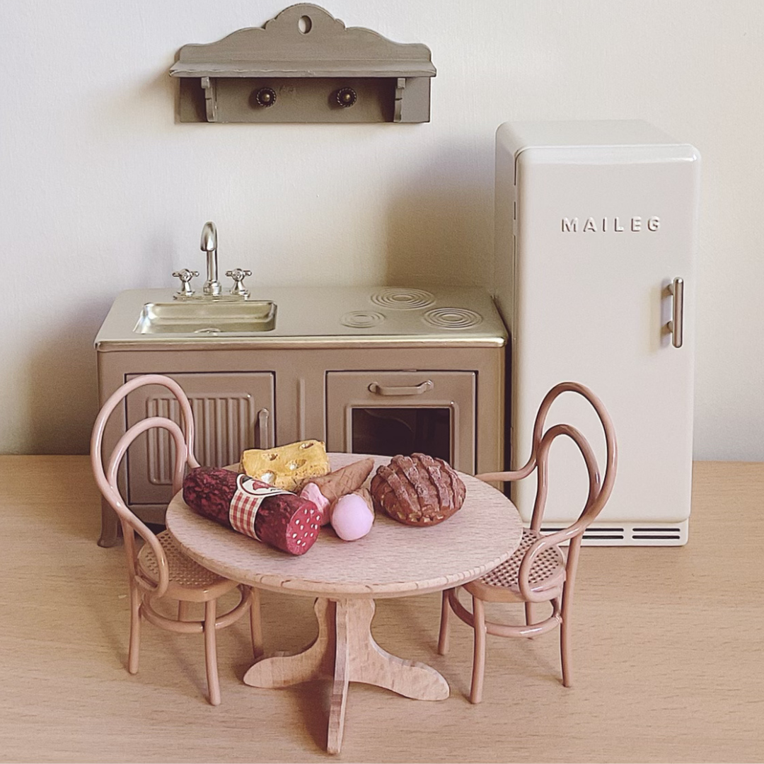 Miniature kitchen set with table, chairs, and refrigerator on a wooden floor.