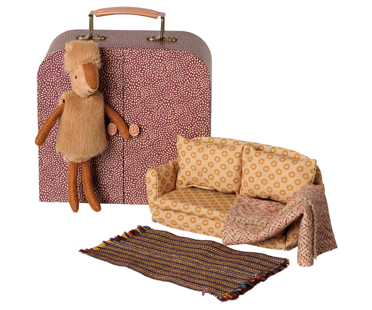 A dollhouse miniature set featuring a patterned suitcase, a small sofa with a throw blanket, and a striped rug, alongside a plush toy sheep.