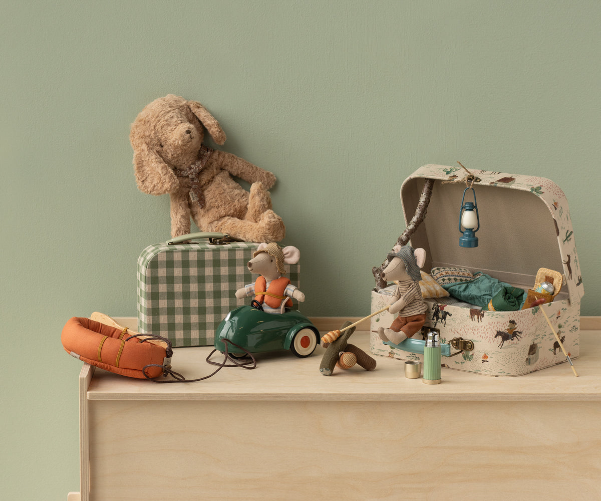 Collection of MAileg toys including a plush dog, toy car, and mice in a suitcase on a wooden surface with a green wall background.