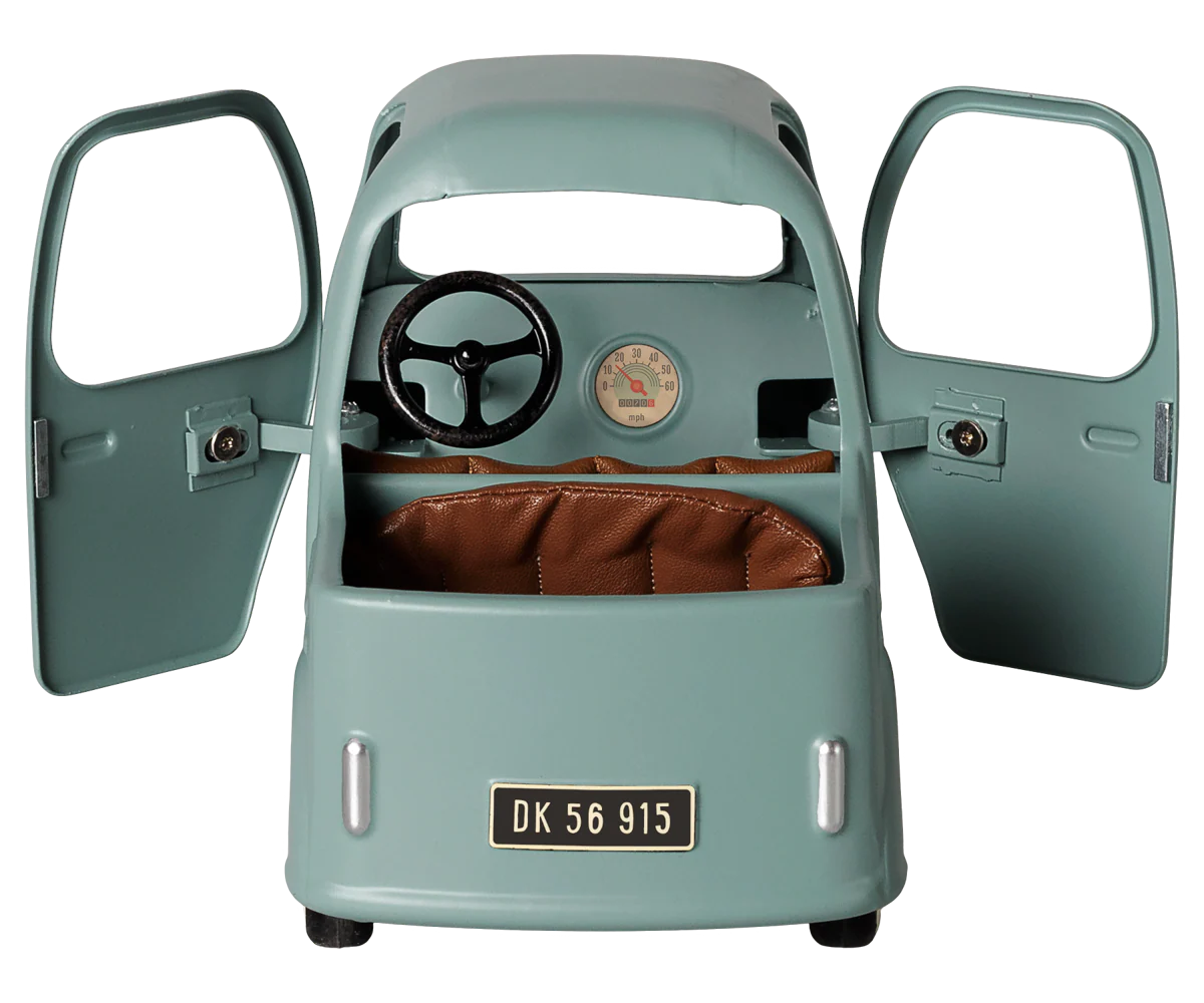 A vintage-style children's ride-on toy car in a muted sage green color, featuring open doors, a black steering wheel, a brown leatherette seat, and a license plate reading 'DK 56 915'.