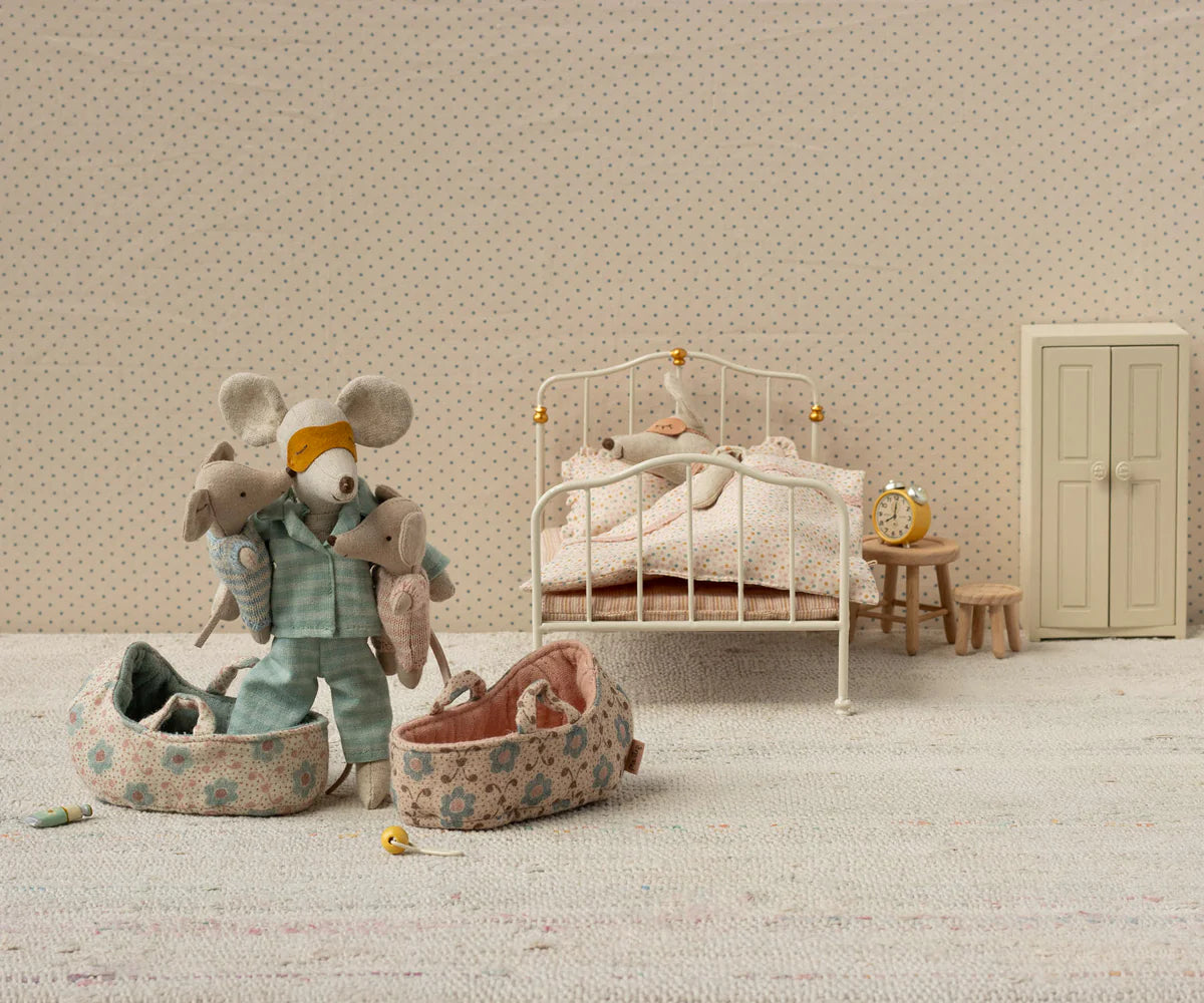 Miniature Maileg bedroom scene with toy baby mice in a carrycot and furniture against a patterned wall.