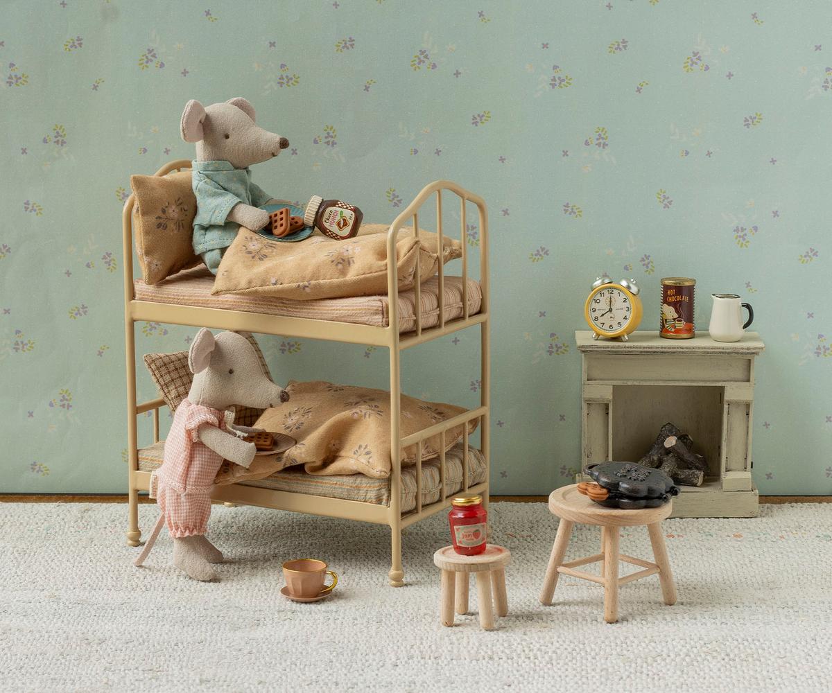 A miniature dollhouse-style room scene with two plush mouse figures arranged on a wooden bunk bed, alongside small furniture pieces, a wallpapered wall, and a fireplace display.