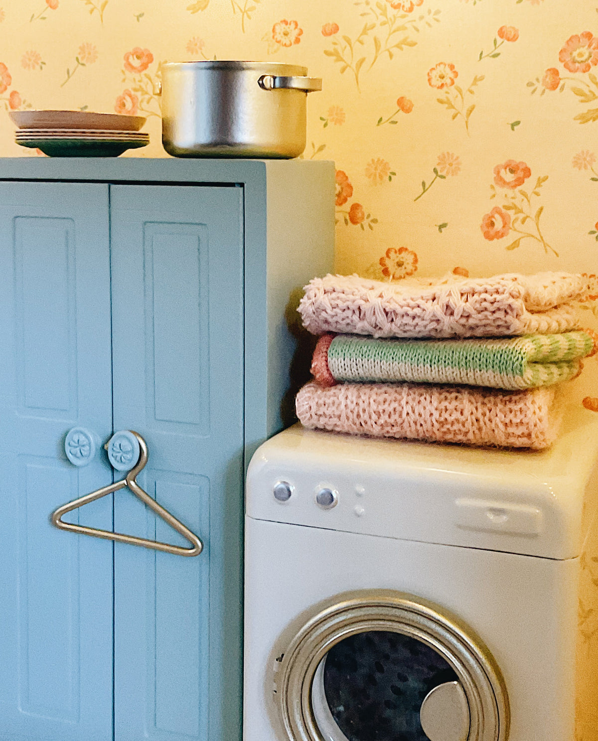 A vintage-style white washing machine with a stack of folded knit blankets in pink, green, and peach on top, next to a light blue cabinet with a metal hanger and a silver pot on the door, set against a yellow floral wallpaper background.