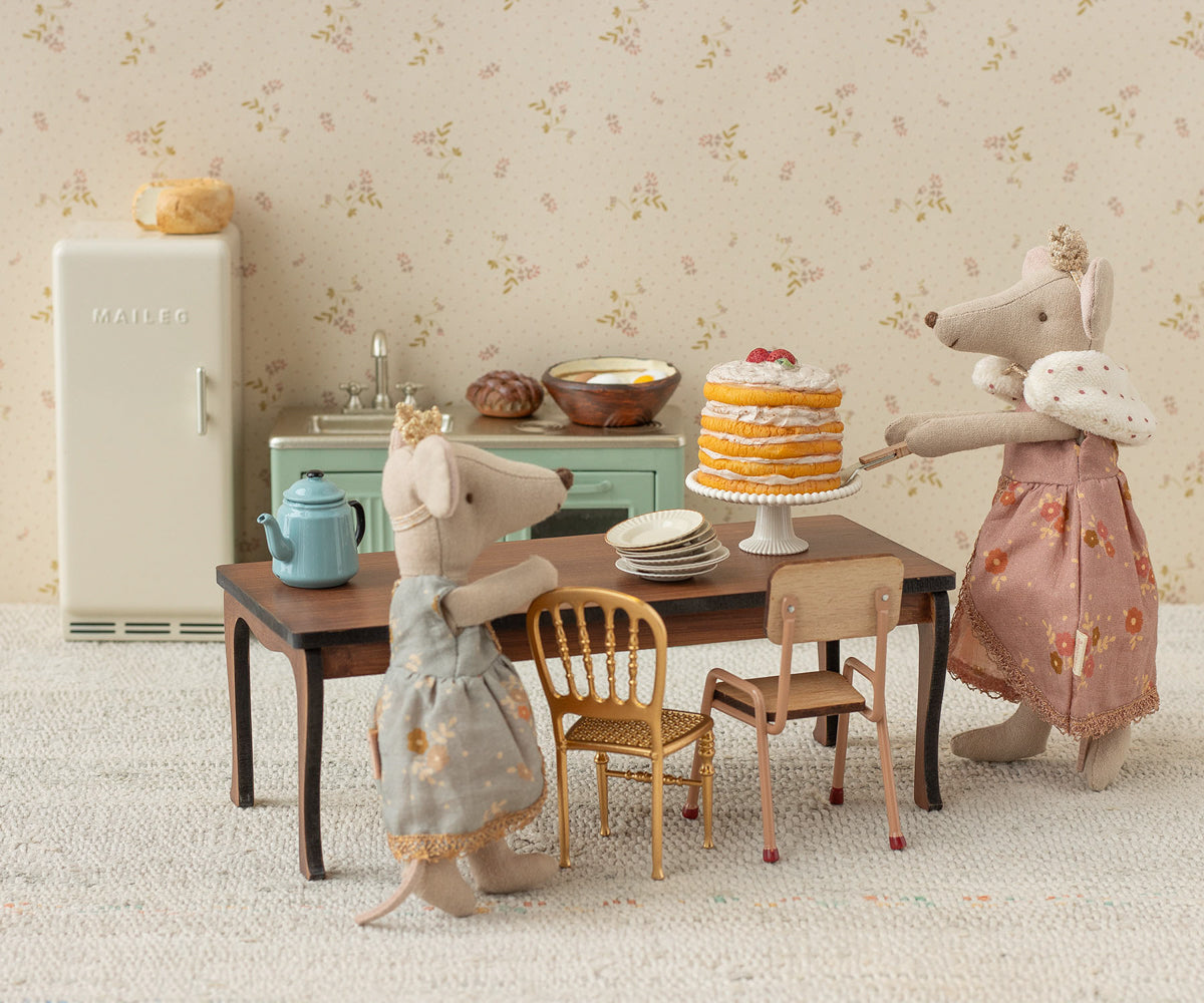 maileg Two toy mice in dresses standing at a table with a cake and tea set, in front of a floral wallpapered wall.