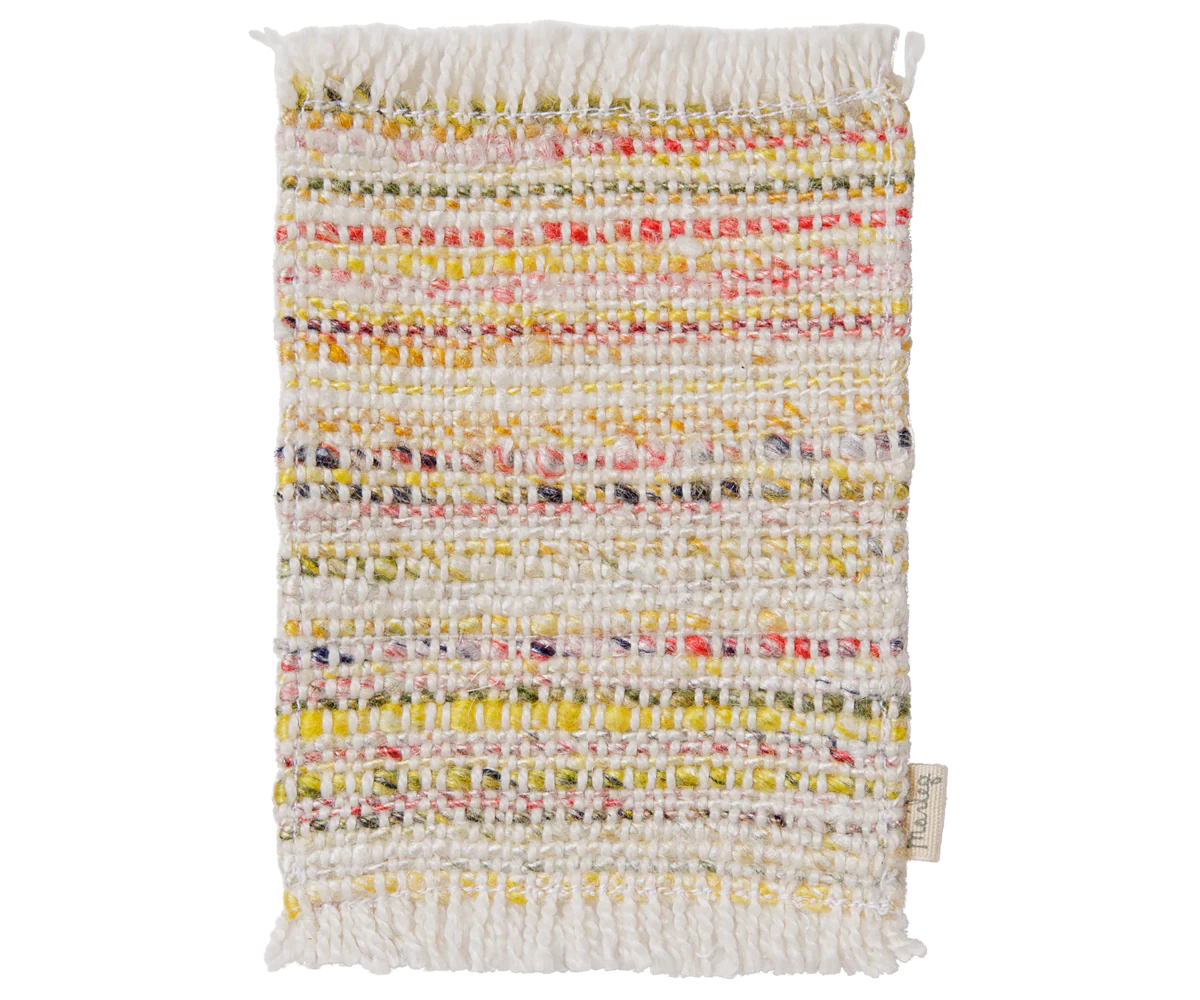 A rectangular woven textile rug or mat with fringed edges, featuring horizontal stripes of multicolored yarns on an off-white base. A small fabric tag is visible at the lower right corner.