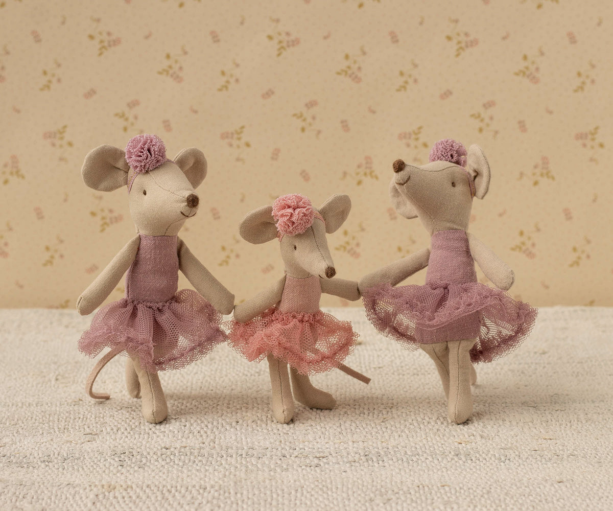 Three toy Maileg mice in ballerina dresses standing on a textured surface with a floral-patterned wall in the background.