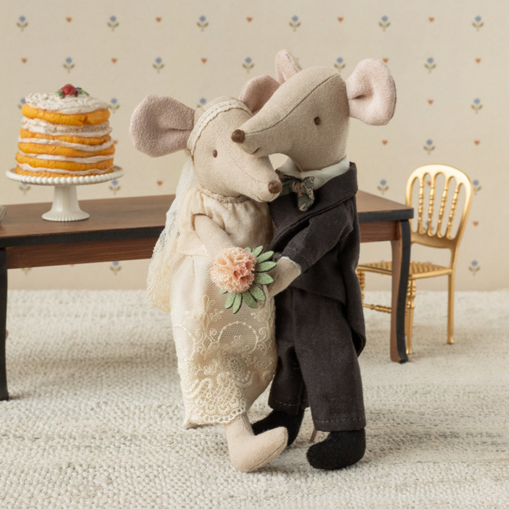 Maileg wedding couple holding hands dressed in wedding outfits in front of a big table with a wedding cake on top