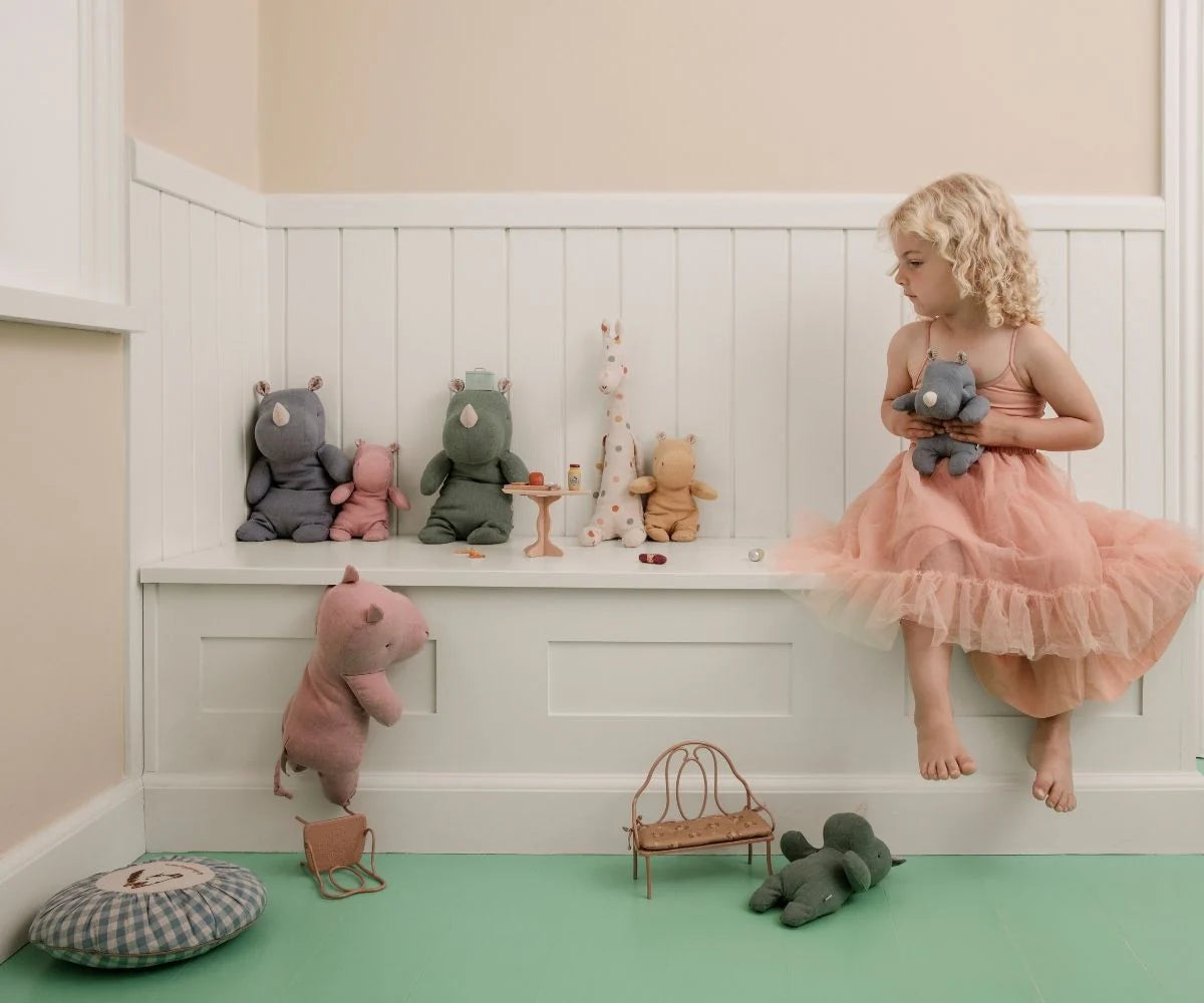 Child in a pink dress sitting on a white shelf with stuffed animals in a room with green floor and beige walls.