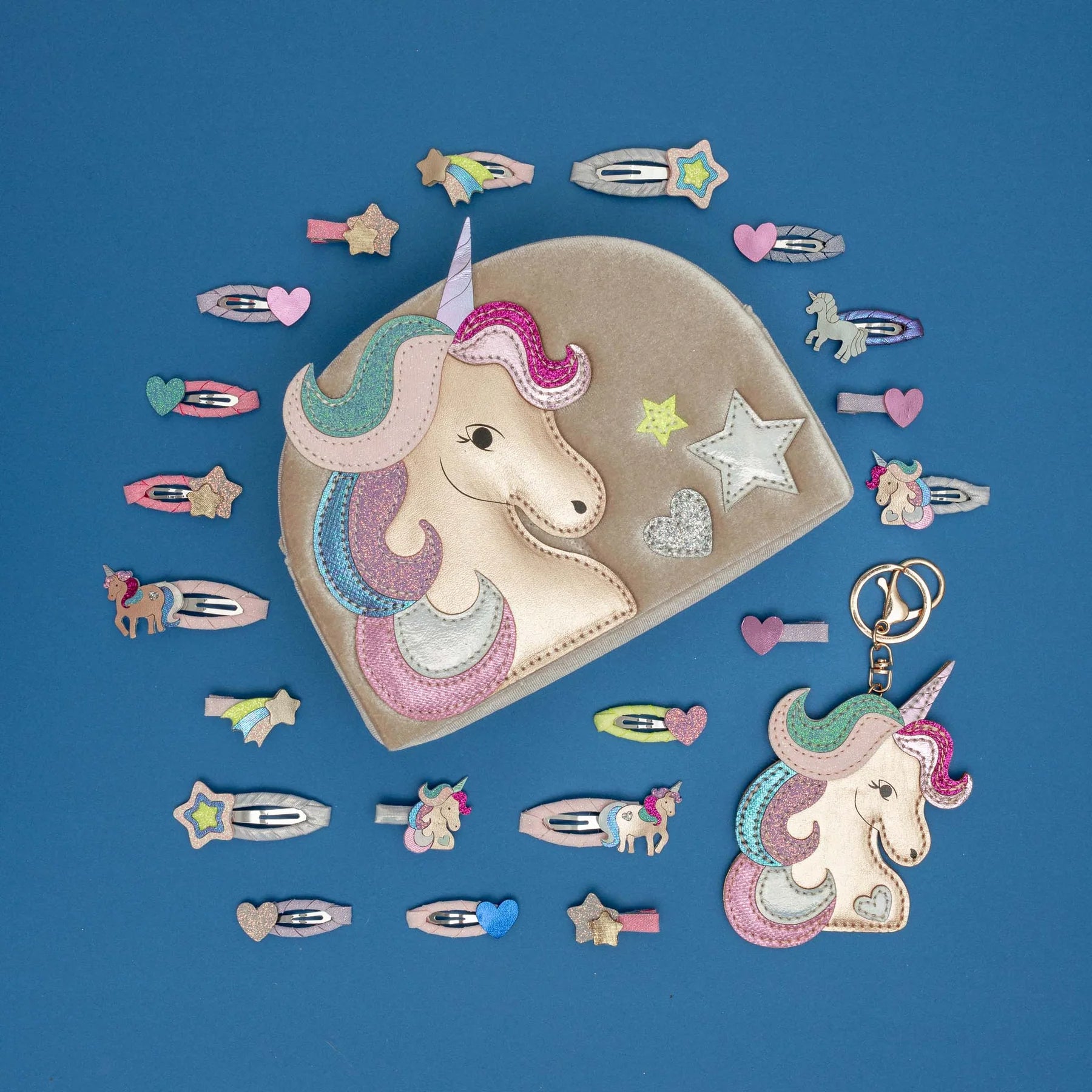 A unicorn-themed fabric hair accessory set arranged on a blue background, including a large beige pouch with a unicorn face applique, several matching hair clips, and a small matching charm with key ring.