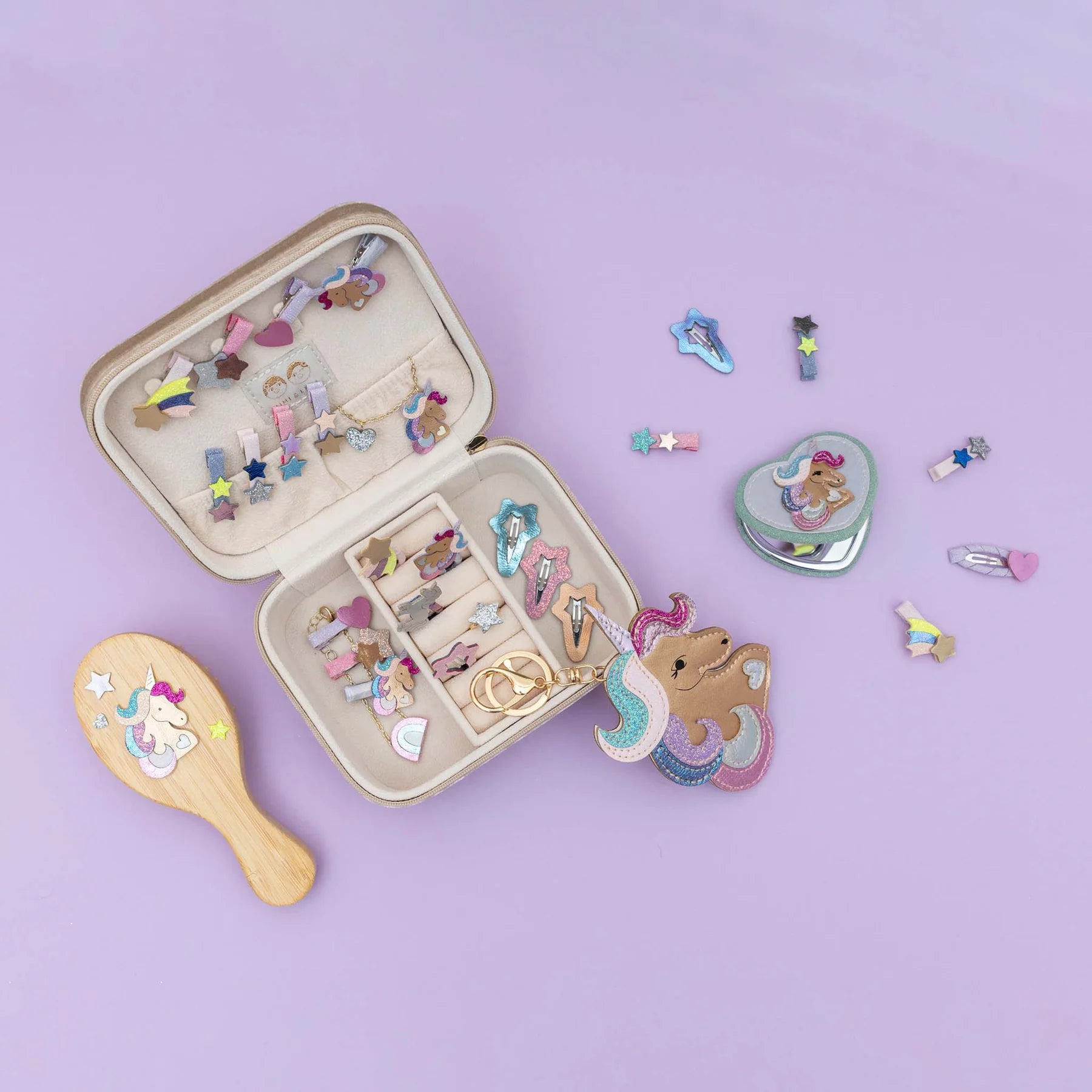 A children's unicorn-themed jewelry organizer set displayed on a lavender background, featuring an open beige case filled with colorful hair accessories, a wooden hairbrush, a heart-shaped compact mirror, and several loose unicorn and star clips.