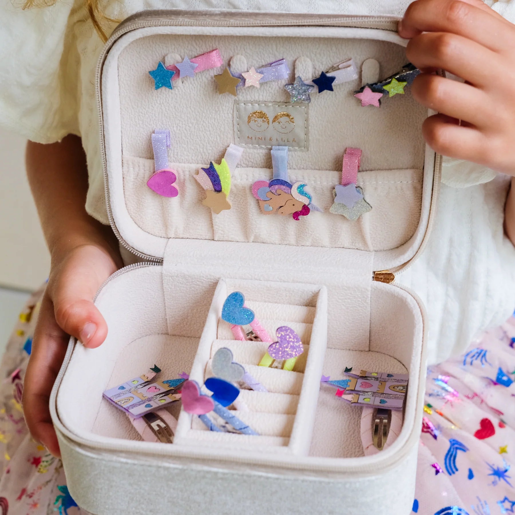 A child's open light gray fabric jewelry organizer case, shown being held by hands. The case is open to reveal multiple compartments holding colorful hair clips and small accessories.