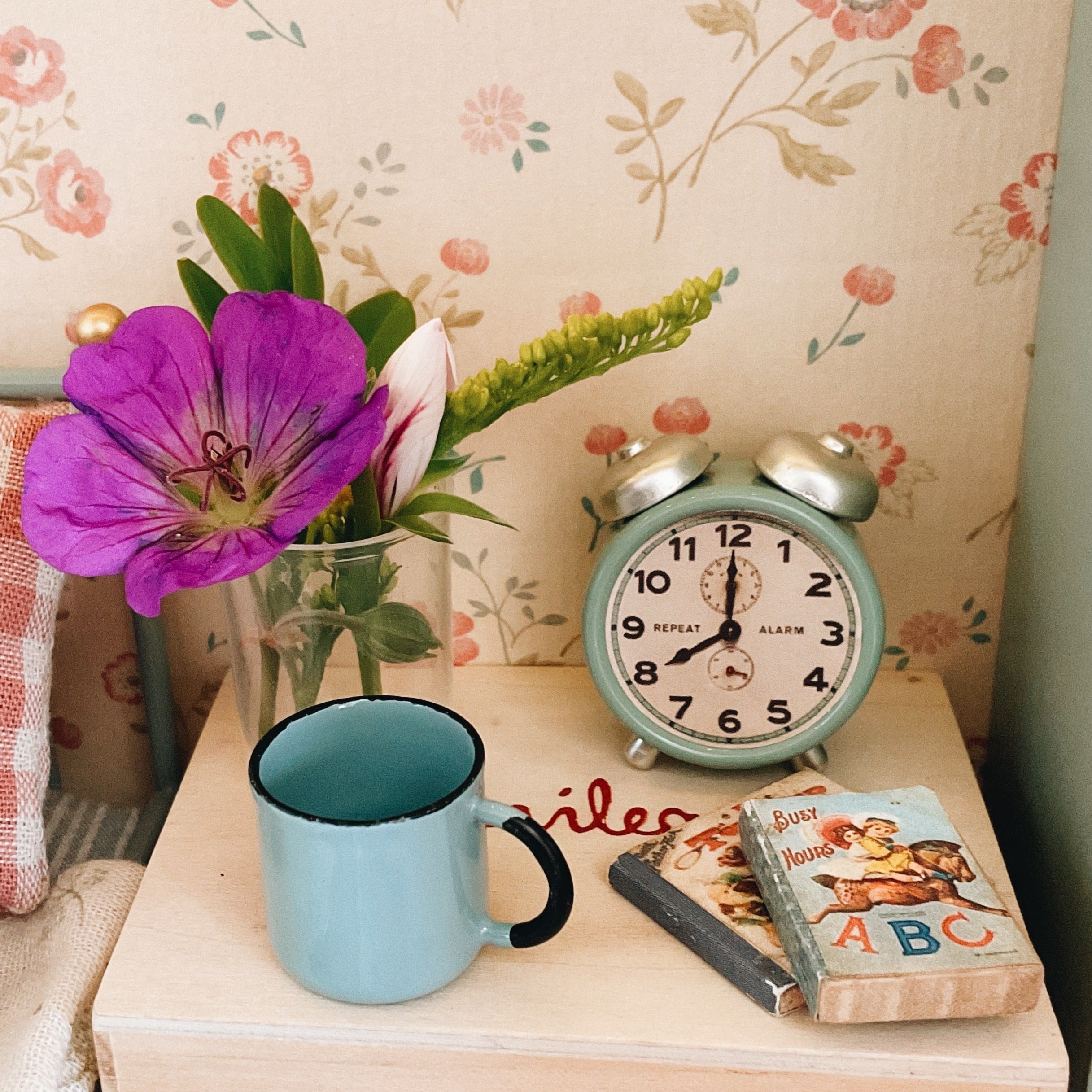 A styled tabletop scene with a light blue enamel mug, a mint green vintage twin-bell alarm clock, a small book, a wooden block, and a glass vase holding a bright purple flower and green stems, set against floral wallpaper.