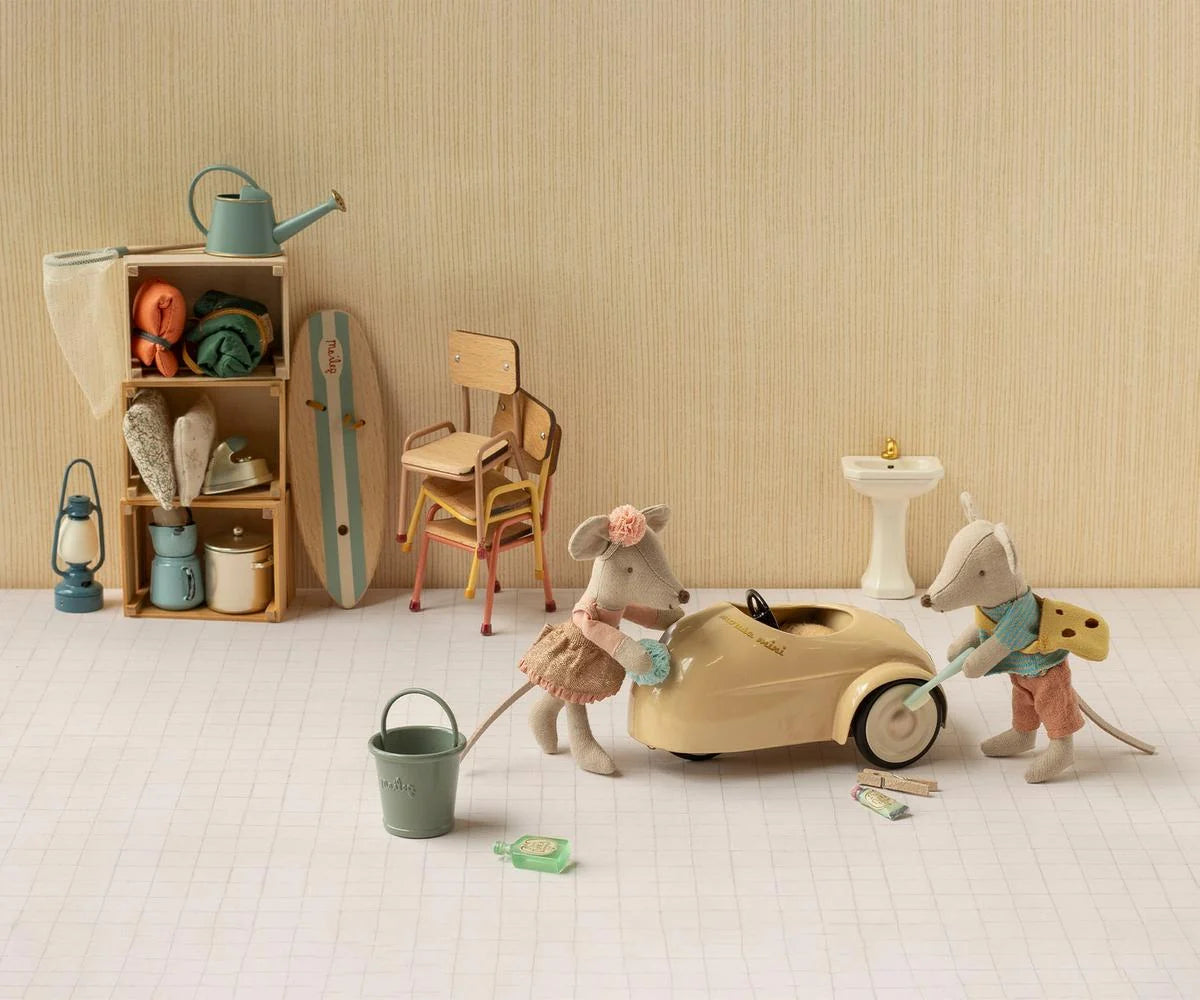 A styled miniature toy scene featuring two mouse figurines playing with a beige toy car, alongside a small bucket, lanterns, a chair, shelving, and other vintage-inspired accessories against a cream striped backdrop.