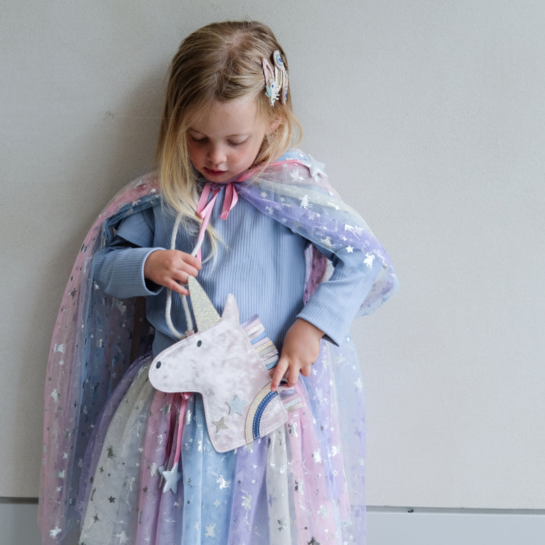 Child wearing a unicorn-patterned cape holding a unicorn plush handbag against a plain background