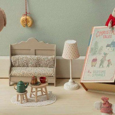 A styled collection of plush toys and miniature accessories arranged on a light wood floor against a pale green wall. The set includes a large pinkish-beige pig plush wearing a red-and-white gingham scarf, a gray mouse plush, a small red-and-white dressed doll, a miniature lamp, chair, and framed picture, plus tiny scattered gold coins.