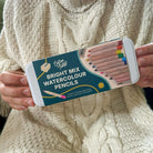 Person holding a set of Cotton Twist Bright Mix Watercolour Pencils against a textured beige background.