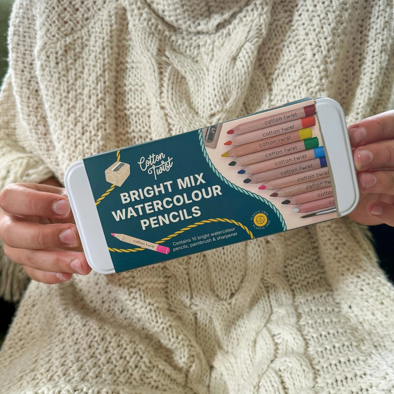Person holding a set of Cotton Twist Bright Mix Watercolour Pencils against a textured beige background.