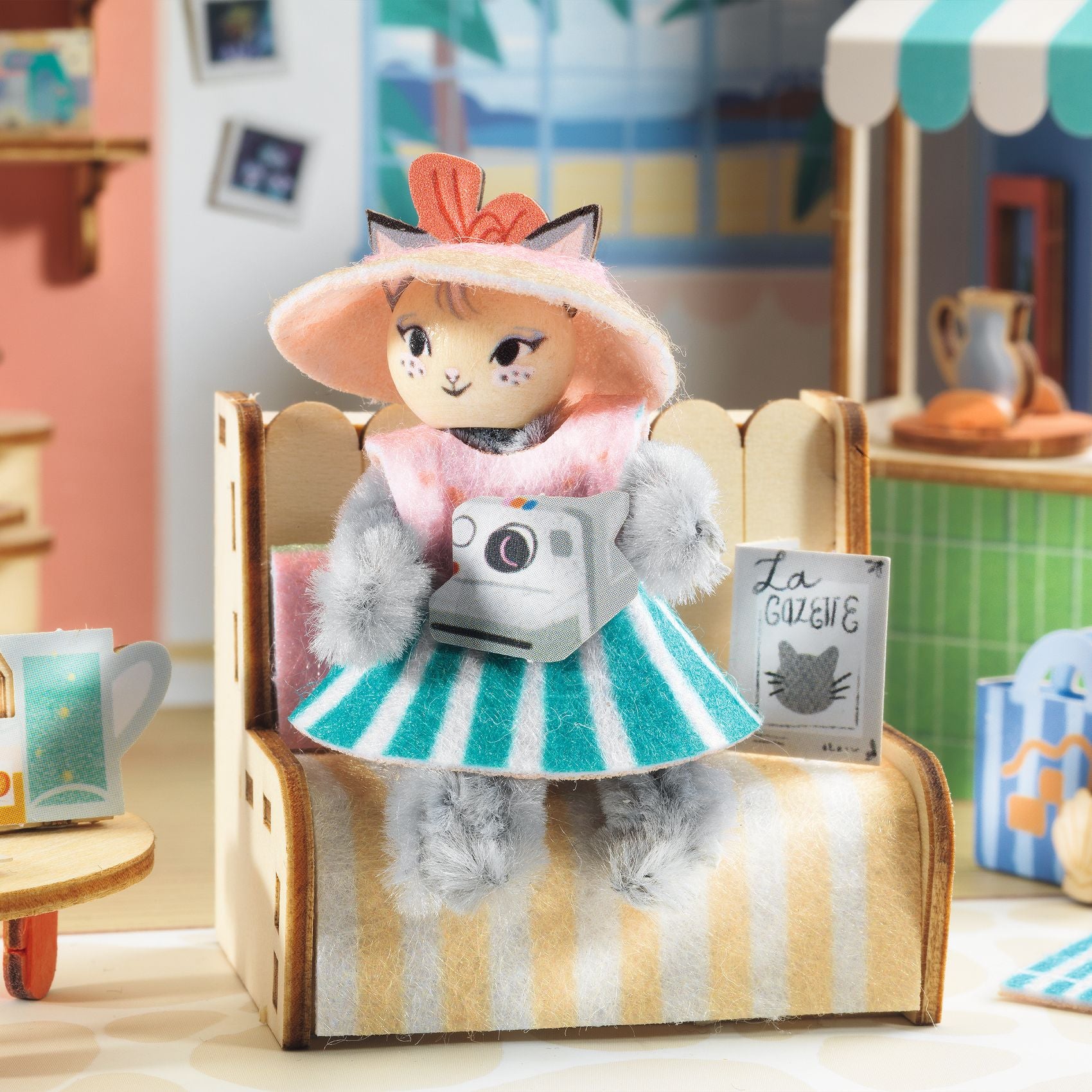 Toy cat in a dress with a camera, sitting on a striped chair in a toy room setting.