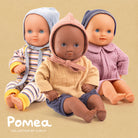 Three dolls with different skin tones wearing colorful outfits on a beige background, with 'Pomea' branding.