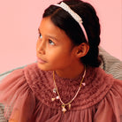 Young girl wearing a ballerina necklace, pink dress with ruffled collar and headband against a pink background