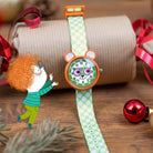 Children's watch with a cartoon character face, green strap, and orange accents on a wooden surface with decorative elements.