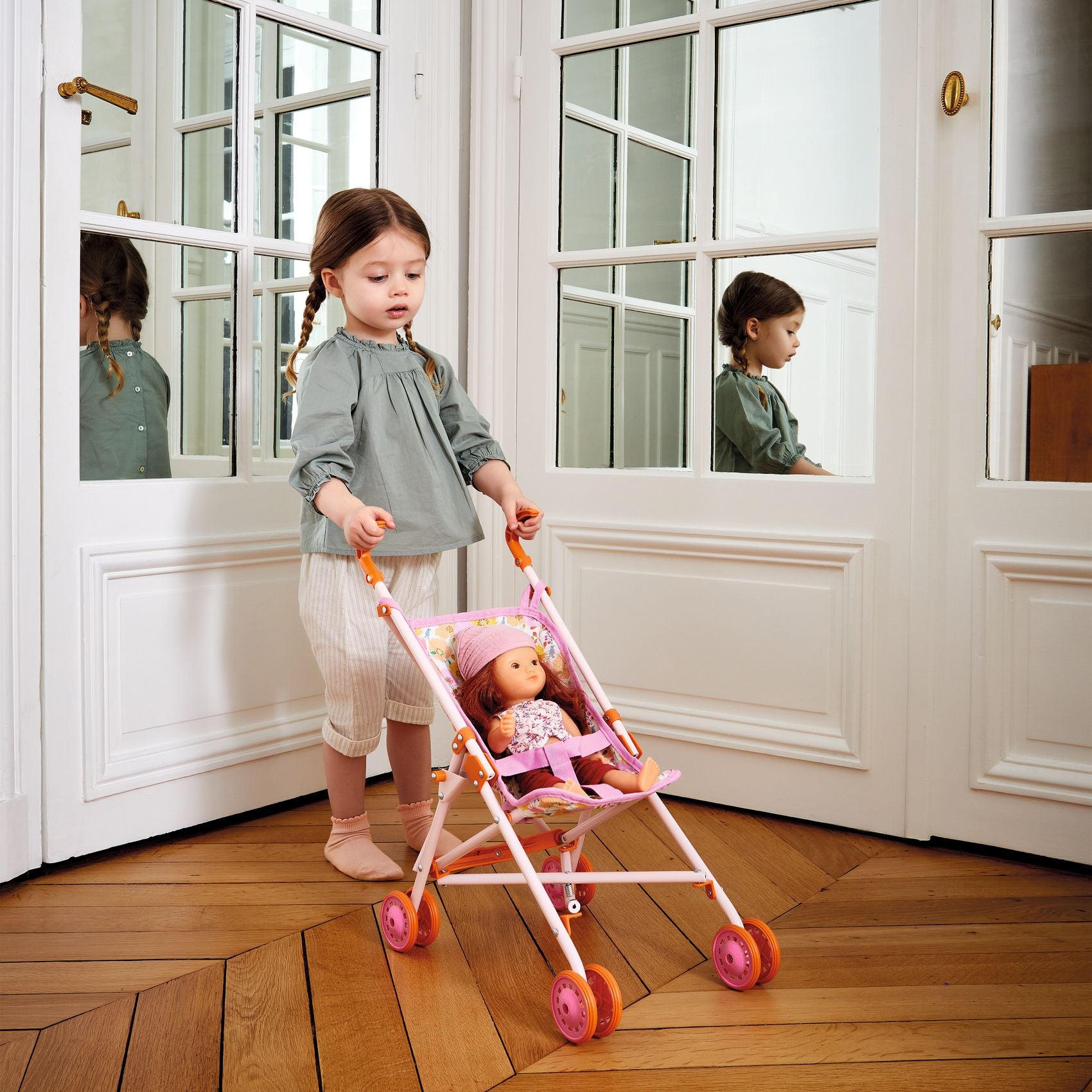 Child pushing a doll in a stroller in a room with large windows.