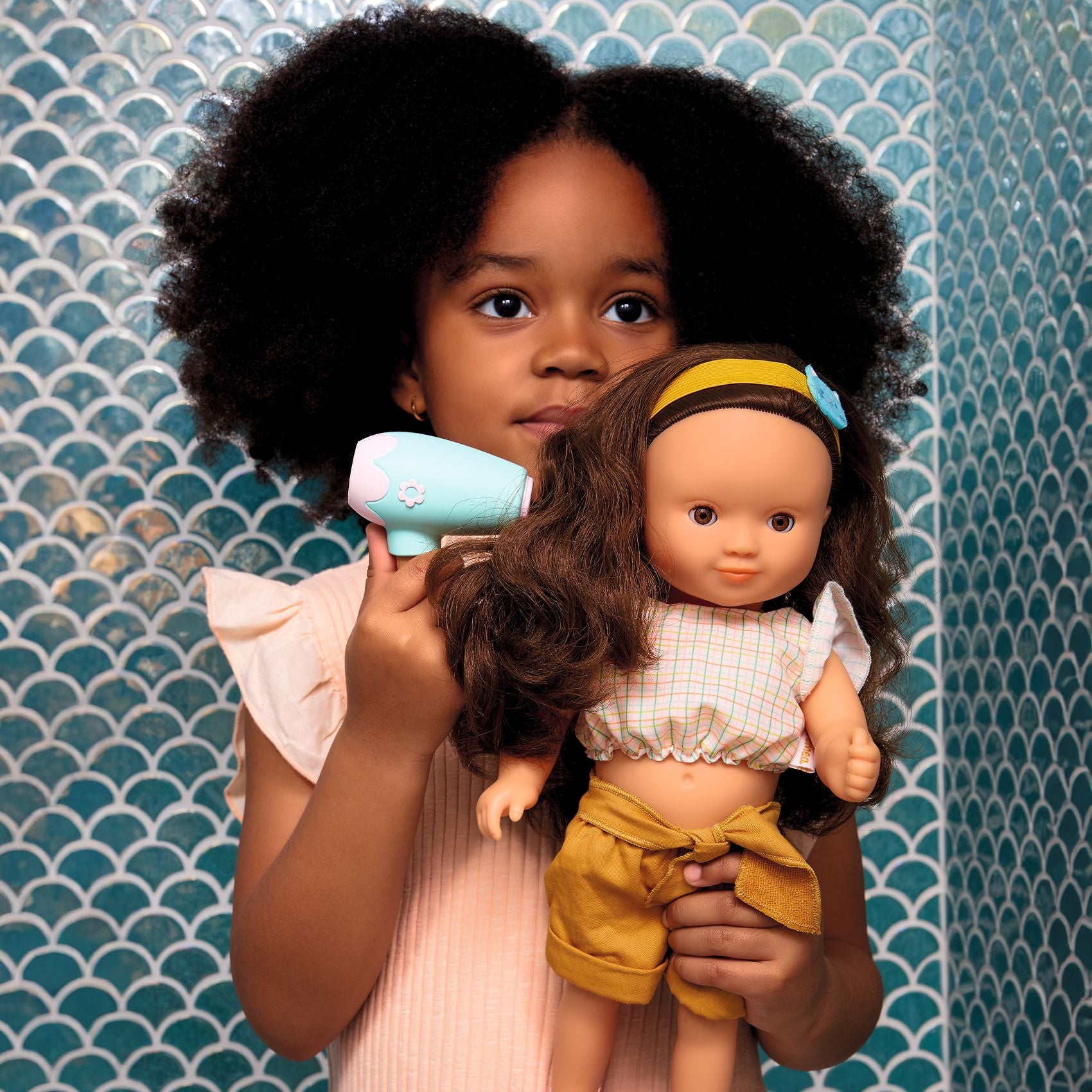 Child holding a doll and a toy hairbrush against a blue patterned wall.