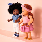 Two dolls, one with dark skin and curly hair, the other with light skin and straight hair, standing against a pink background.