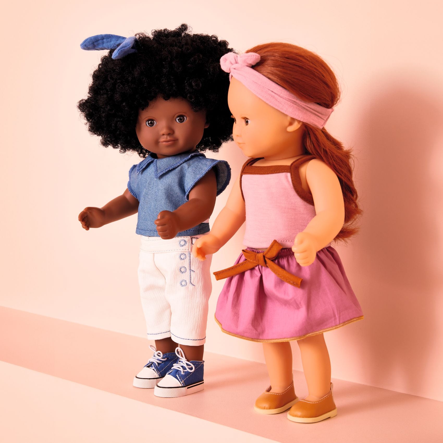 Two dolls, one with dark skin and curly hair, the other with light skin and straight hair, standing against a pink background.