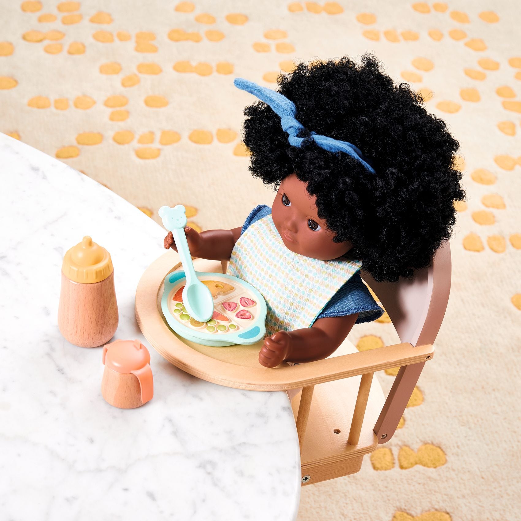 Doll with curly hair sitting at a table with a plate of food and utensils on a patterned background