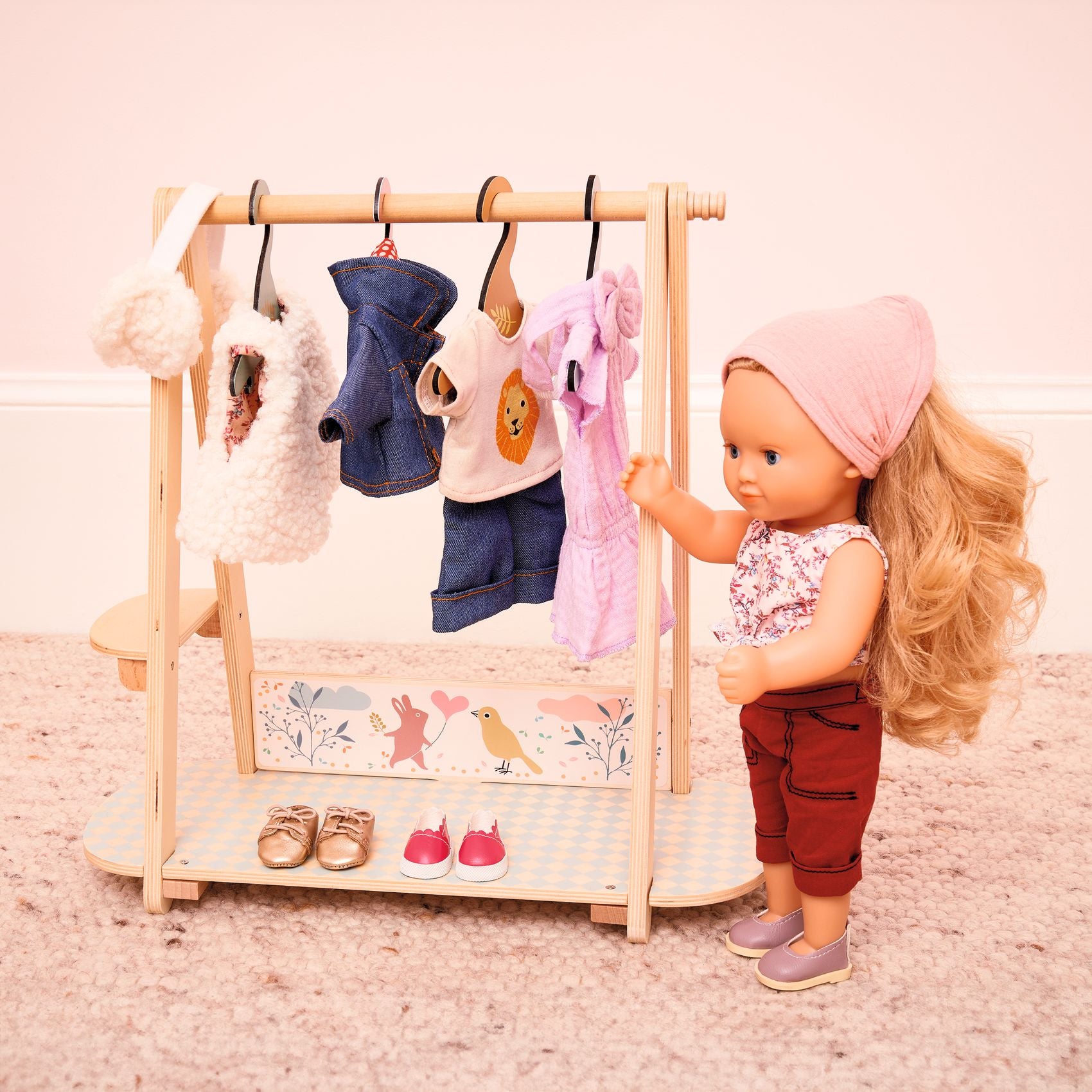 Doll interacting with a wooden clothing rack with hanging clothes and shoes on a pink background