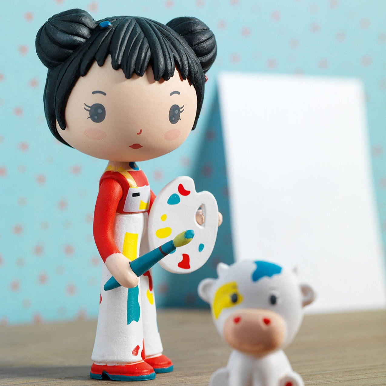 Figurine of a  character with black hair and colorful outfit holding a paintbrush and palette, standing next to a small cow figurine on a wooden surface.