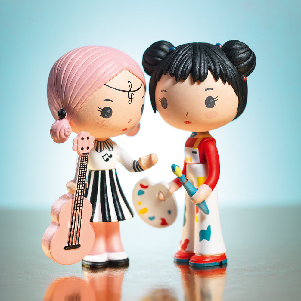 Two stylized figurines, one with pink hair and the other with black hair, holding musical instruments and art supplies on a reflective surface.