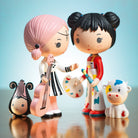 Two figurines, one with pink hair holding a guitar and the other with black hair with painting  accessories, on a reflective surface with a light blue background.