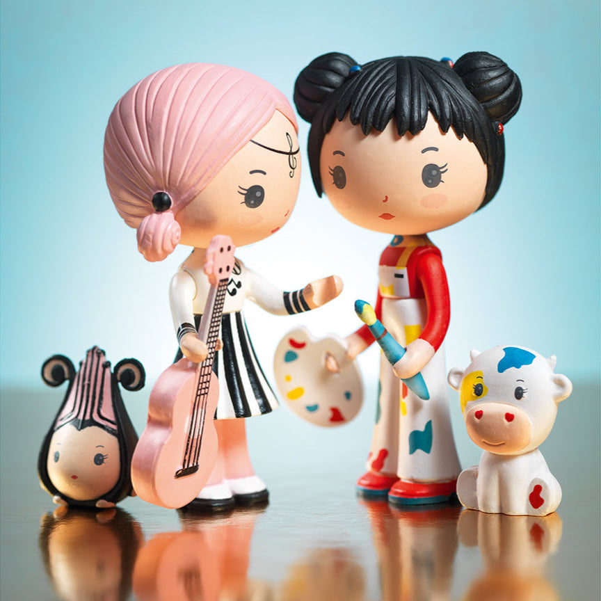 Two figurines, one with pink hair holding a guitar and the other with black hair with painting  accessories, on a reflective surface with a light blue background.