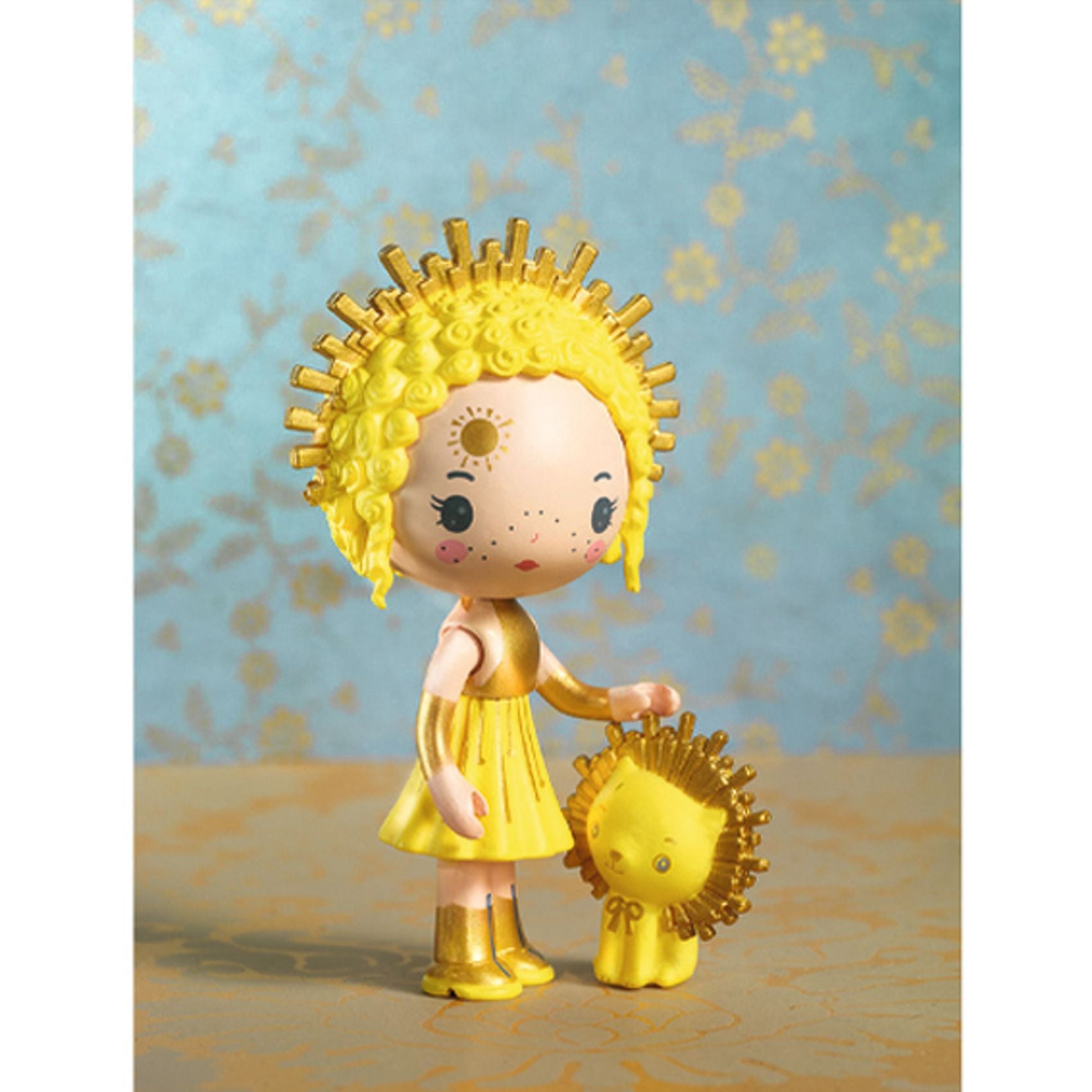 Toy figure of a child with yellow hair and dress standing with a small yellow lion against a floral-patterned background