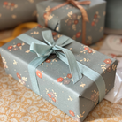 A wrapped gift box in front and a second box behind it, both wrapped in muted sage green paper with a small floral print and tied with ribbons, set on a patterned beige fabric surface.