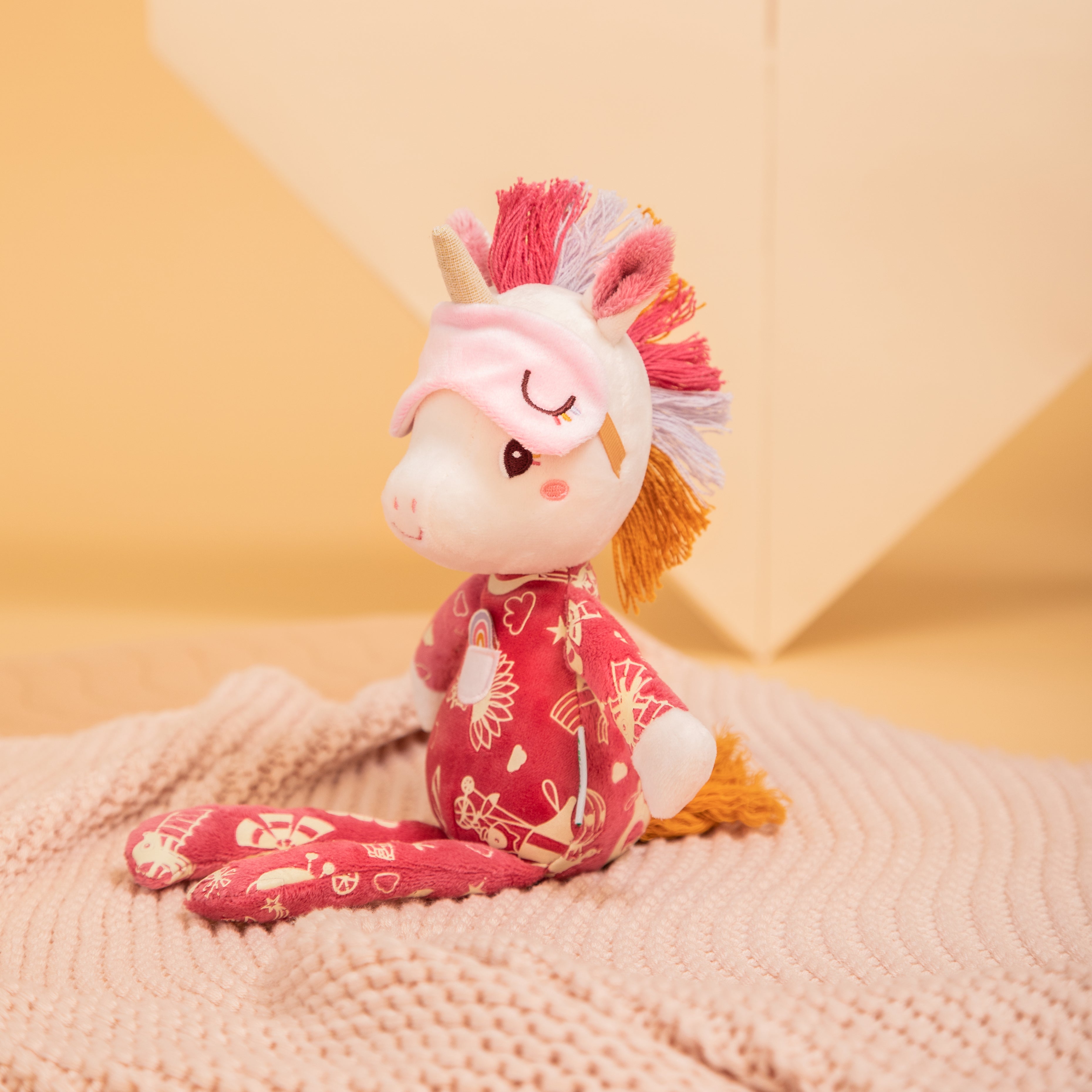 Stuffed unicorn toy with pink mane and red patterned body on a textured surface with a yellow background